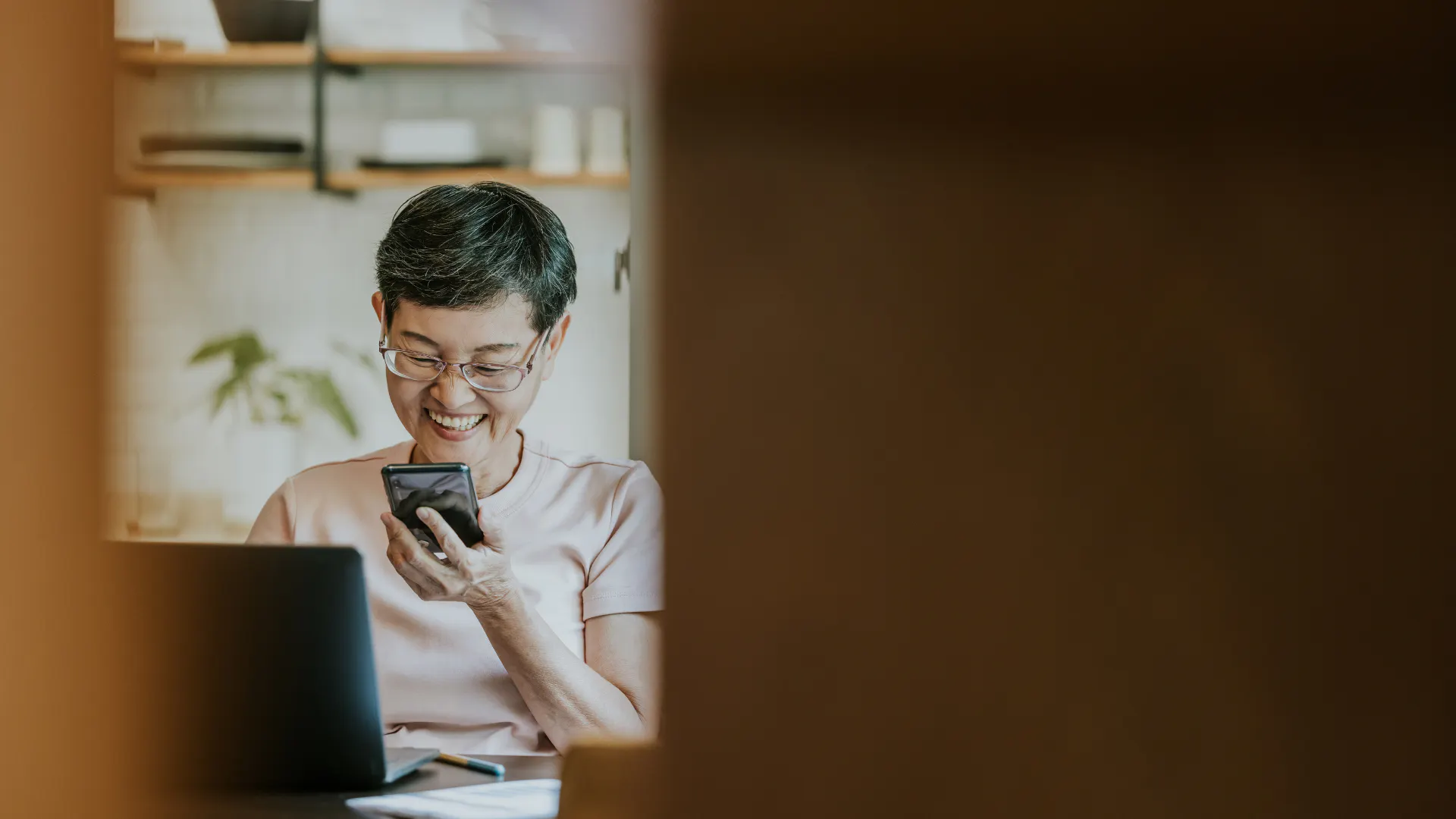 Woman of Asian descent looking at phone and seated in front of laptop in kitchen