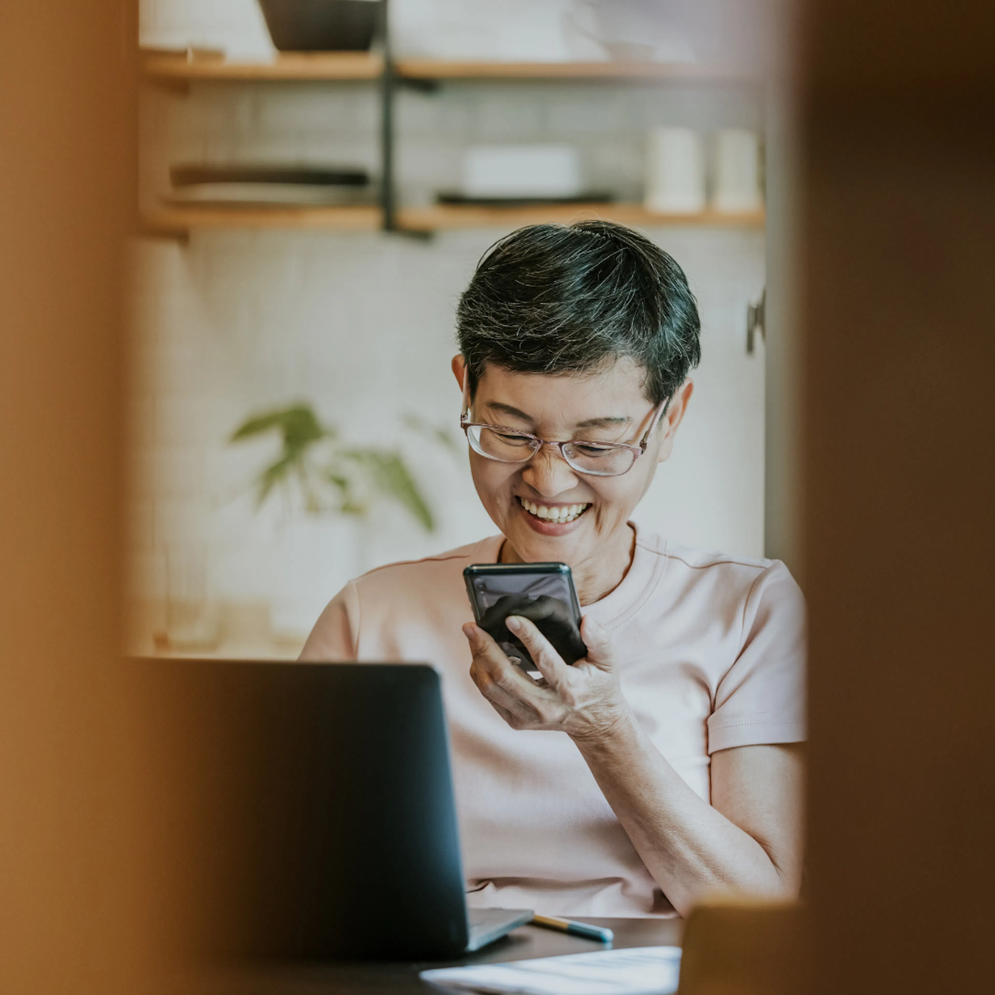 Woman of Asian descent looking at phone and seated in front of laptop in kitchen