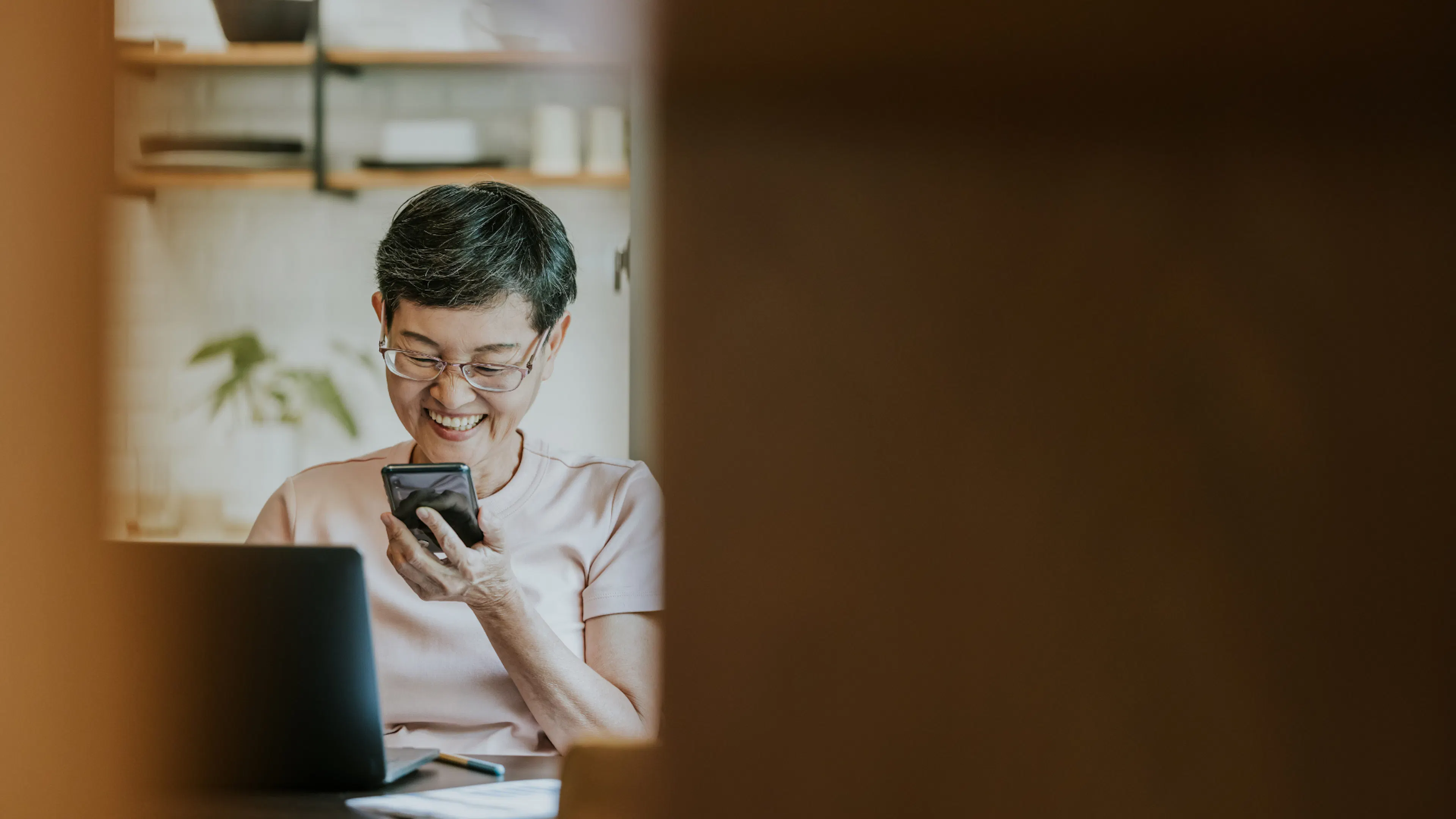 Woman of Asian descent looking at phone and seated in front of laptop in kitchen