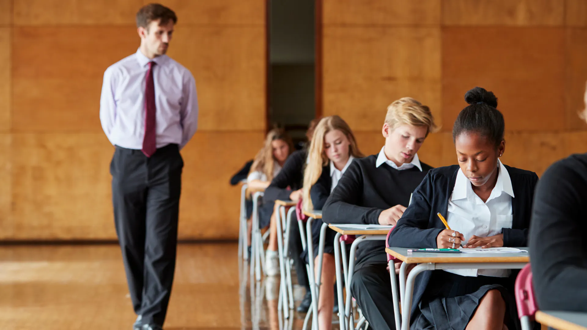 Male exam invigilator walking hall with school children at desks