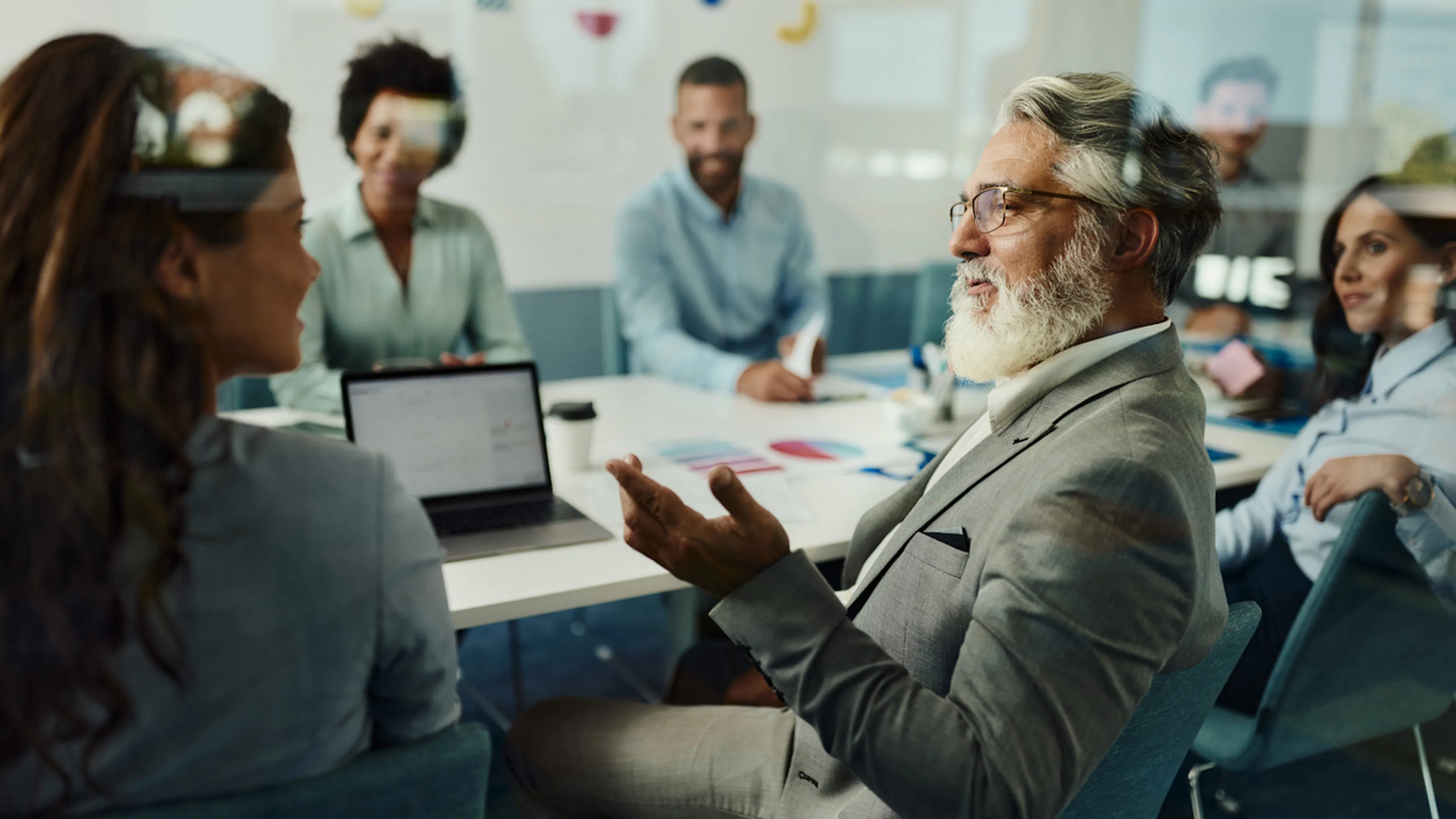 Man in office looking at woman explaining something from laptop, middle aged, glasses, white beard