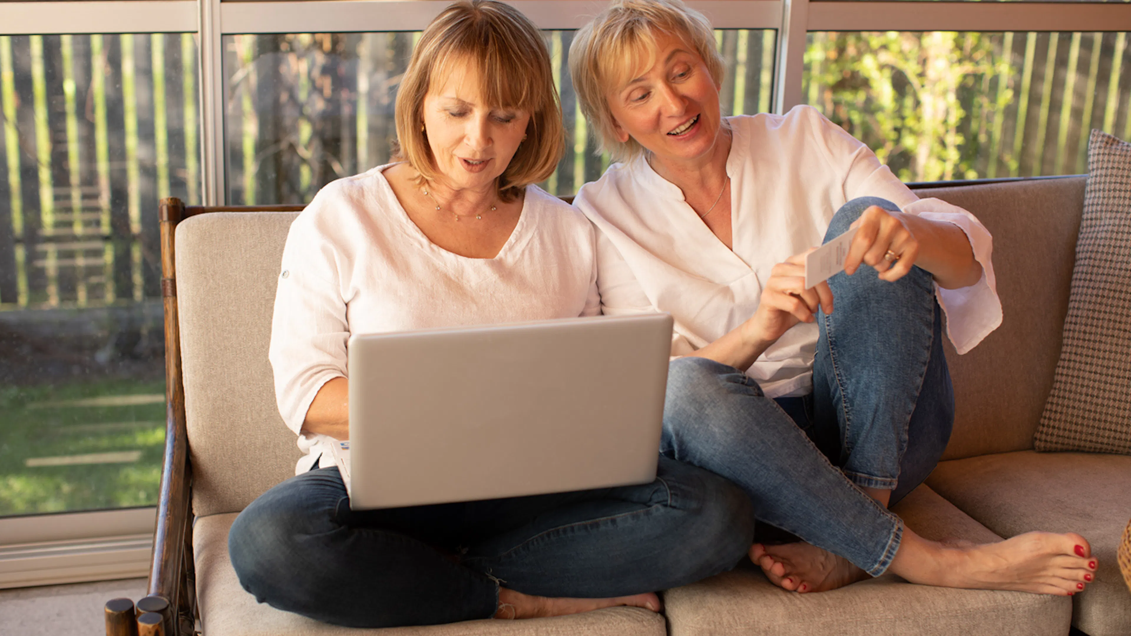 two women middle aged sitting on sofa with laptop wearing jeans