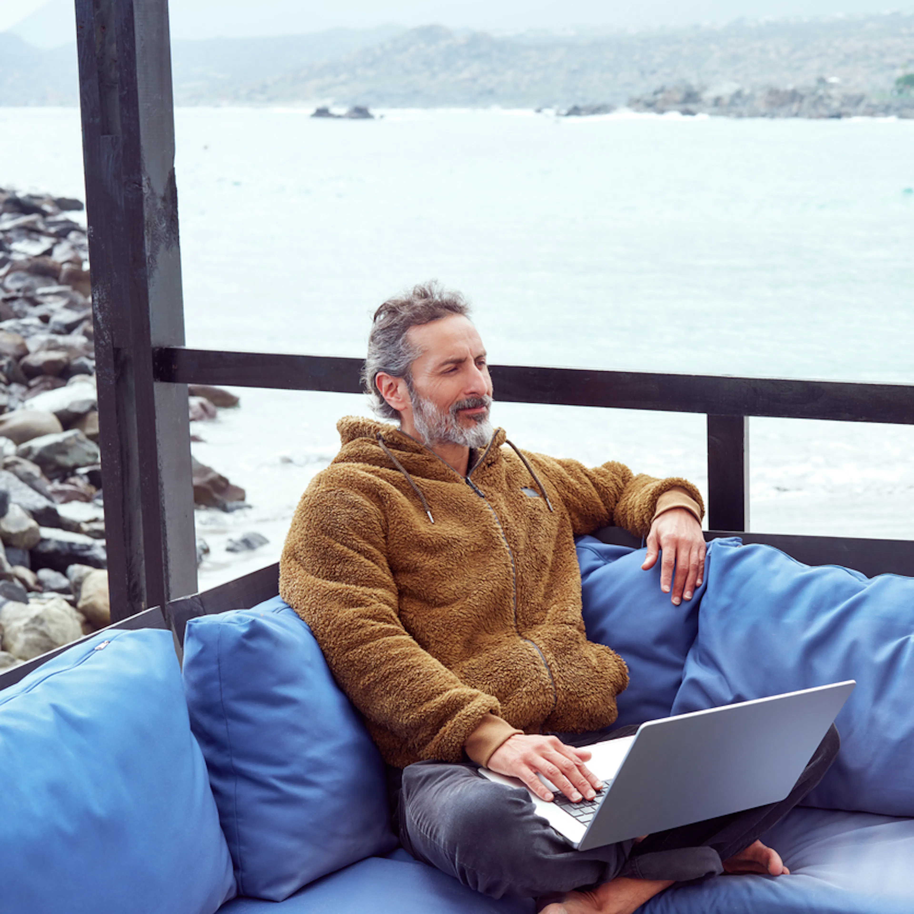 Man sitting on a sofa with a laptop and a guitar with a sea view

