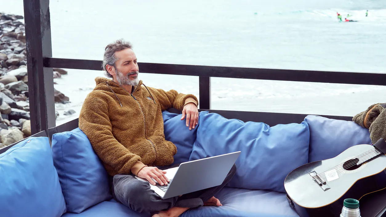 Man sitting on a sofa with a laptop and a guitar with a sea view