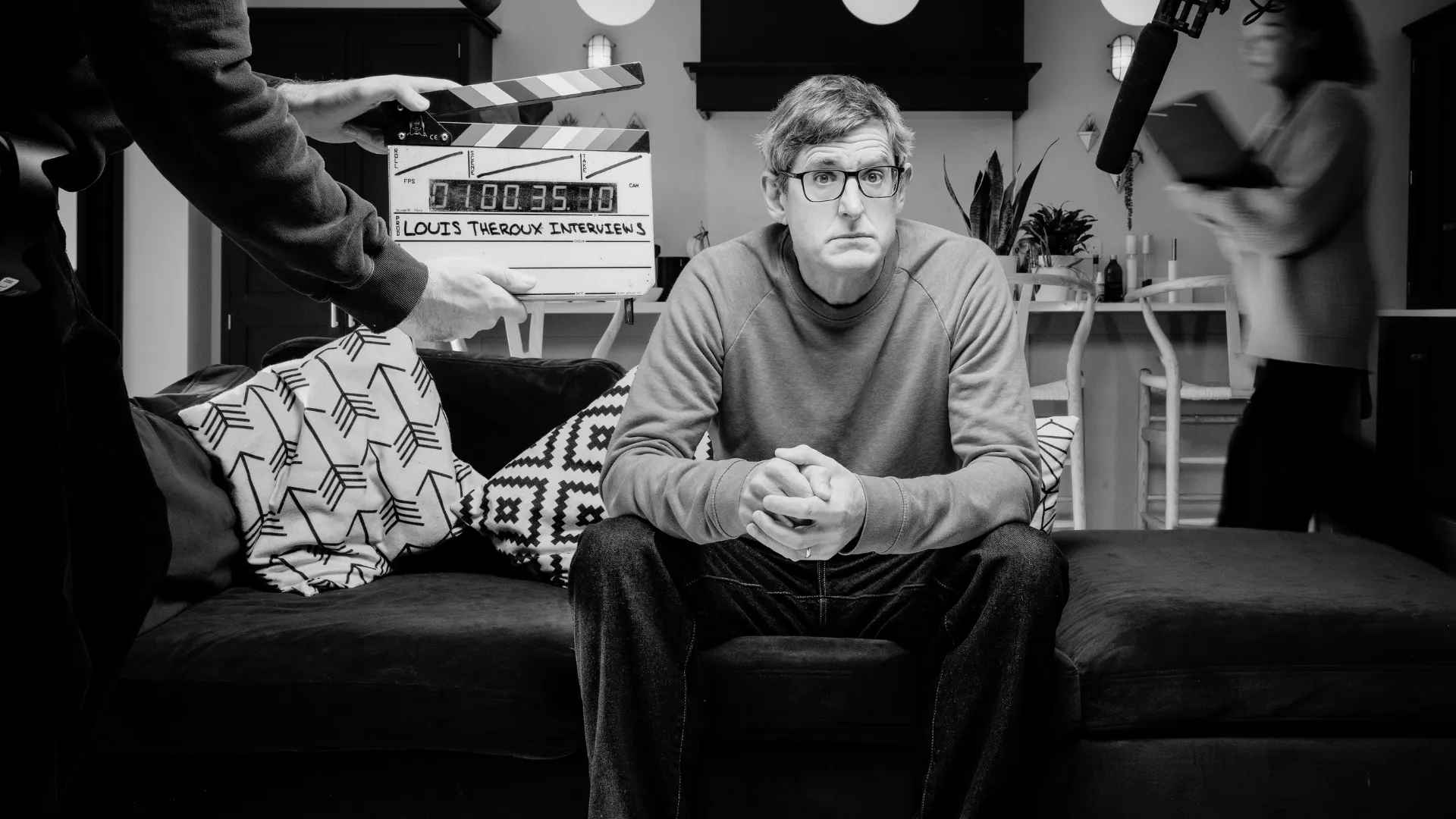 Louis Theroux sat on sofa looking at camera with filming activity is happening around him