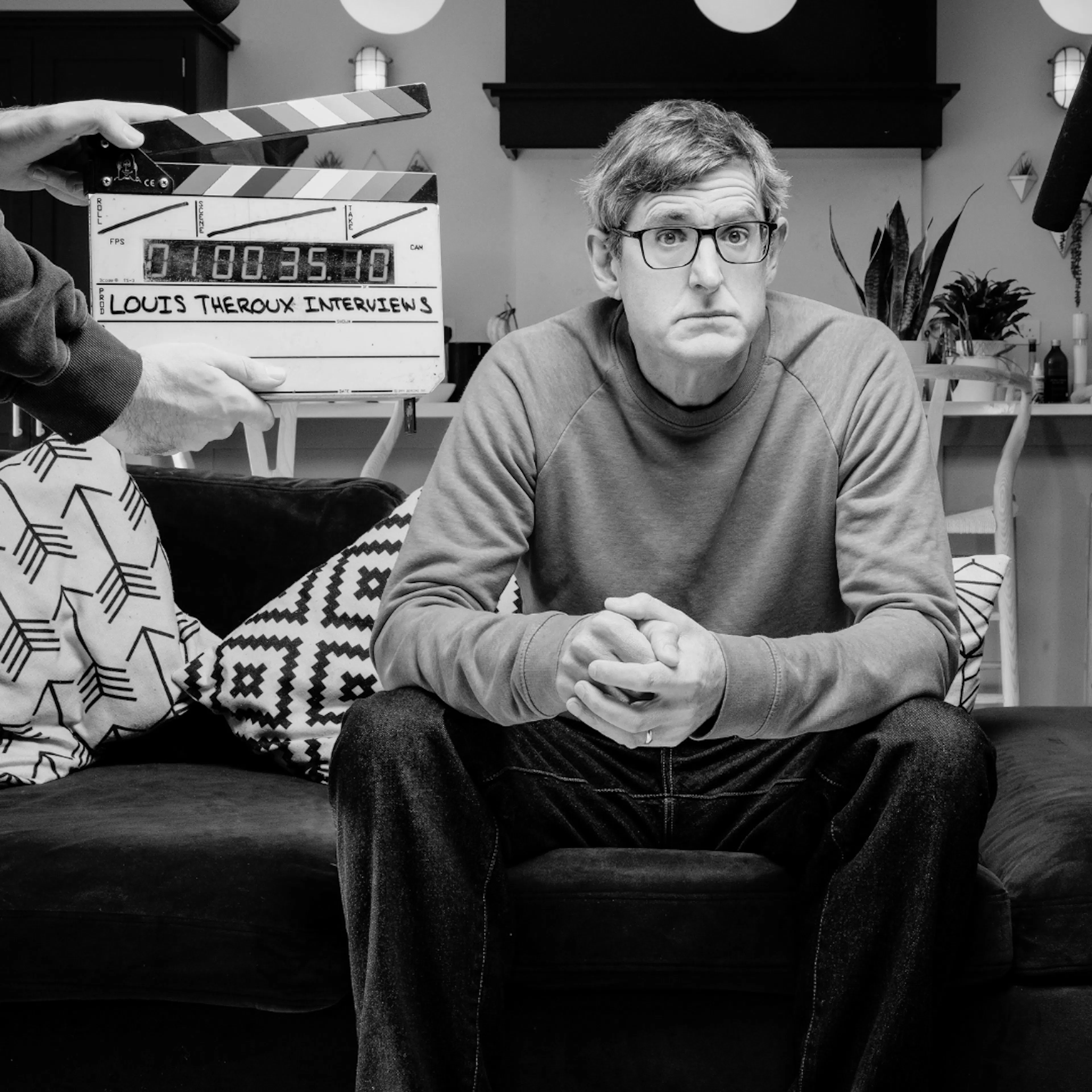 Louis Theroux sat on sofa looking at camera with filming activity is happening around him