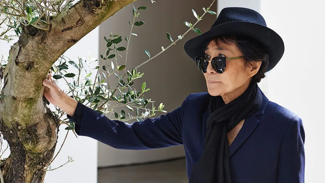 Yoko Ono poses next to a tree in her trademark hat