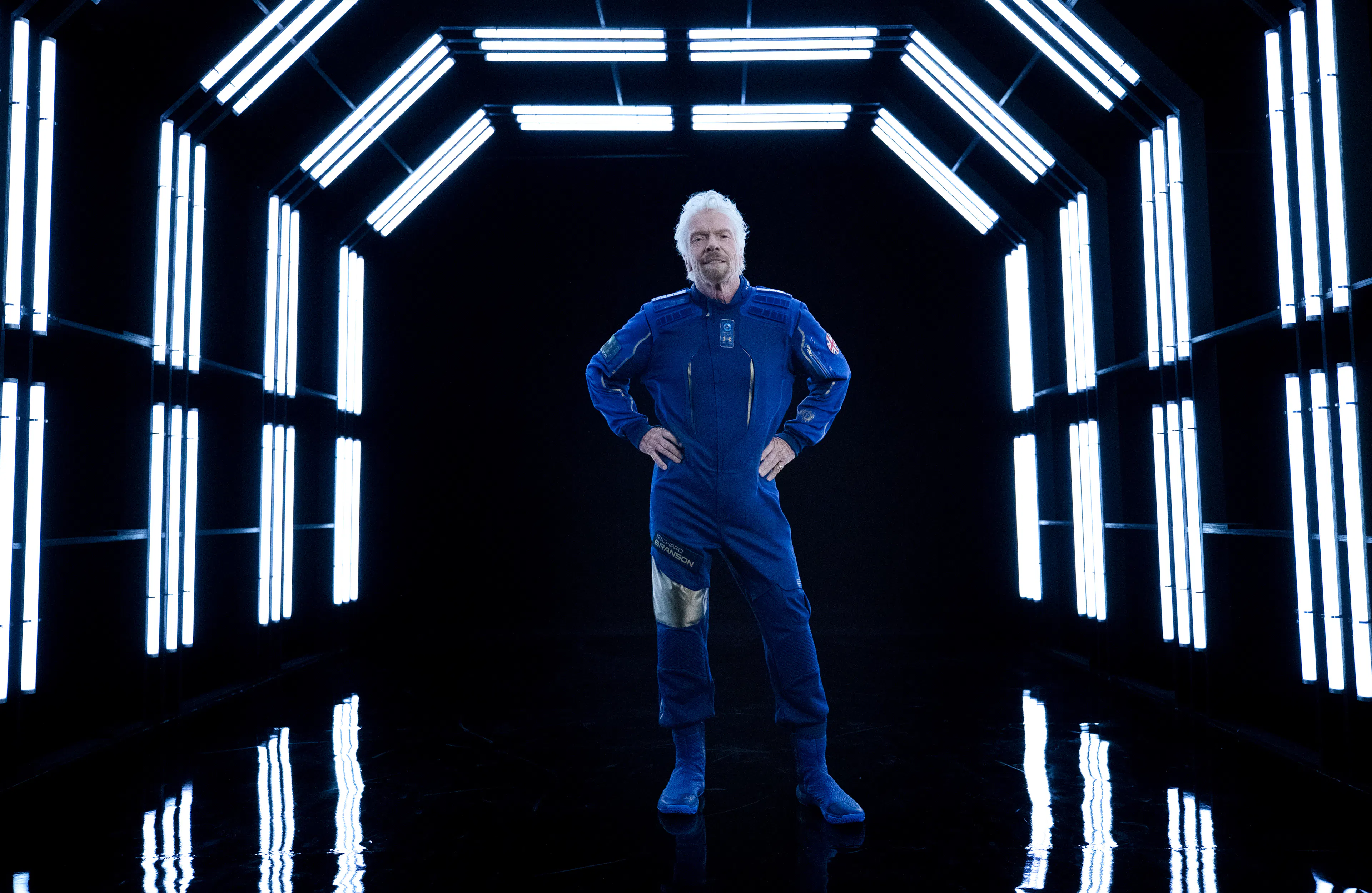 Richard Branson in a black corridor. Wearing a blue flight suit