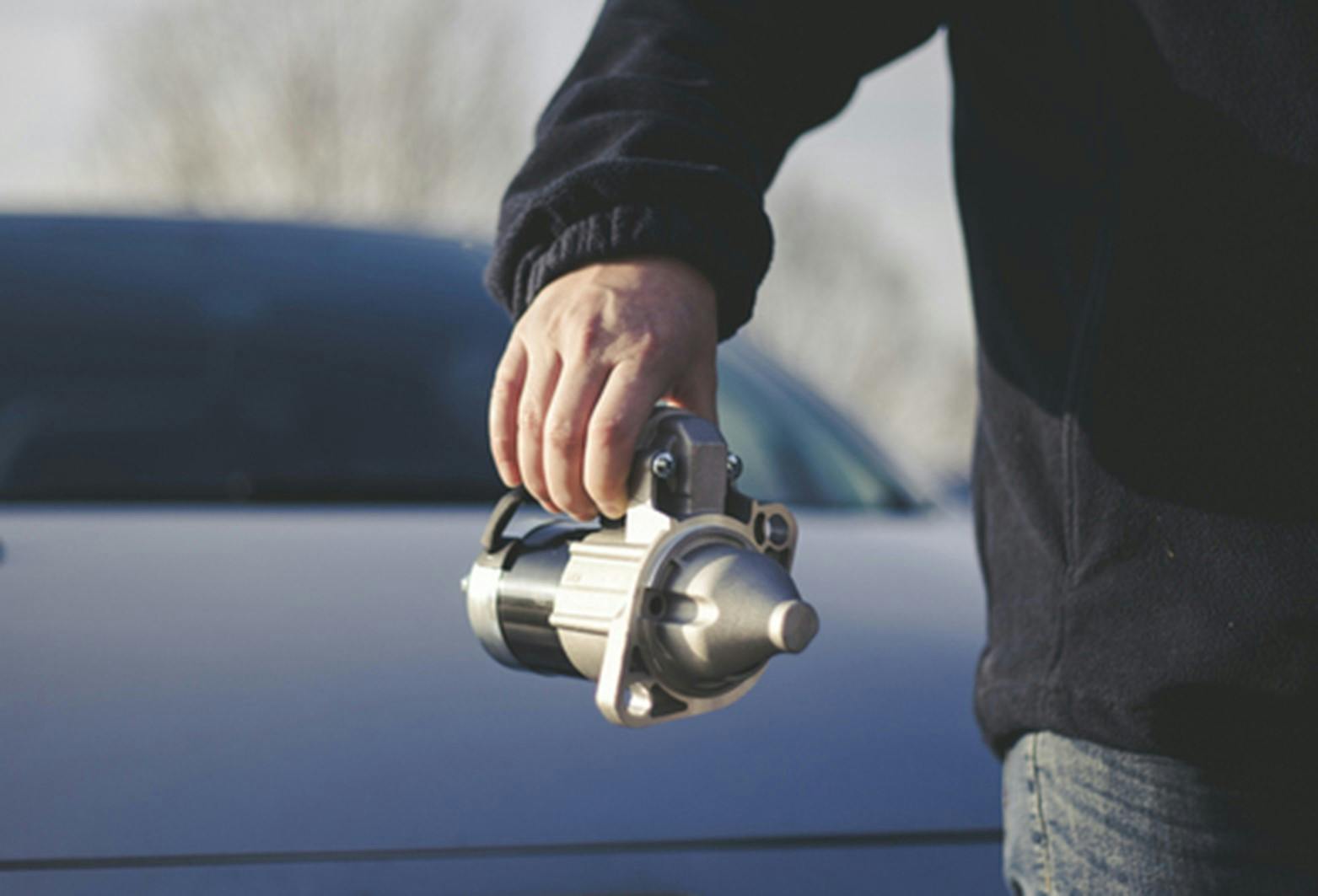 A man holding a starter motor with a car in the background