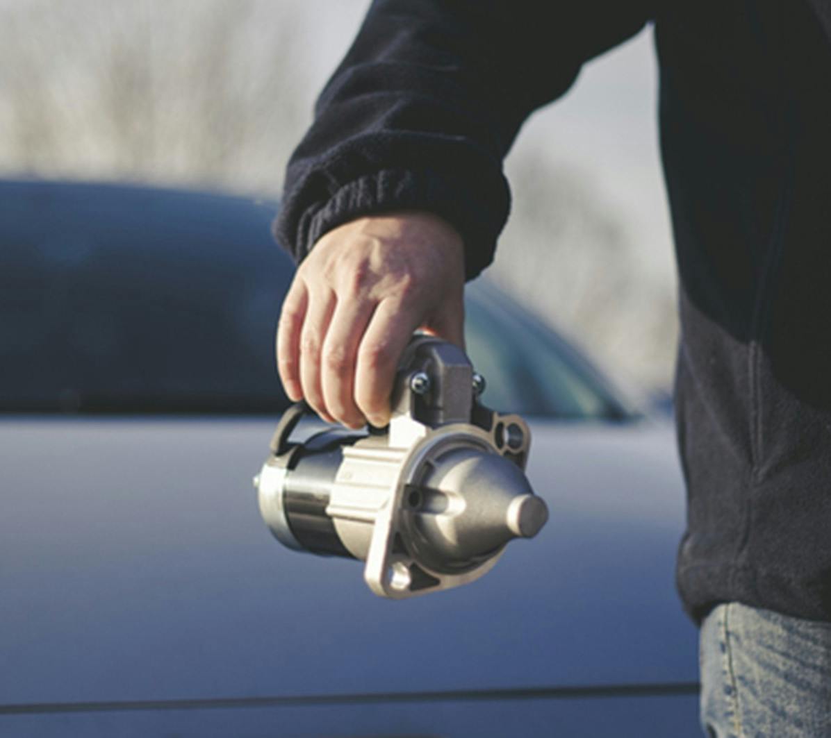A man holding a starter motor with a car in the background