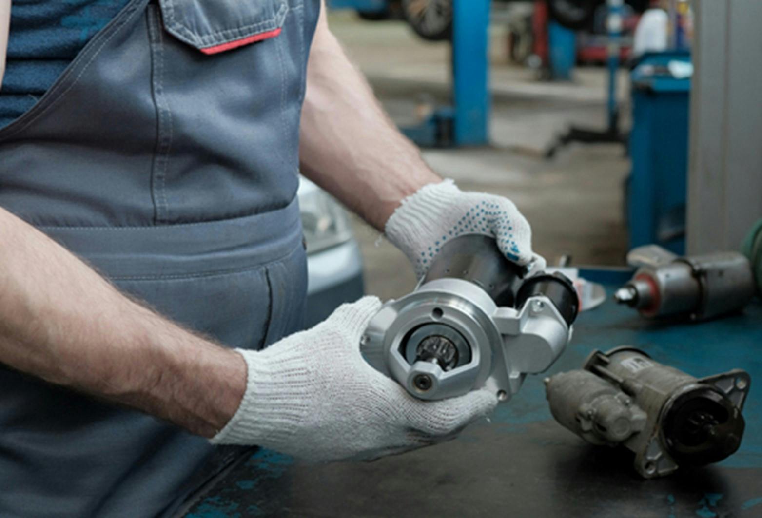 A mechanic wearing gloves holding a new starter motor with the old starter motor sitting on a workbench