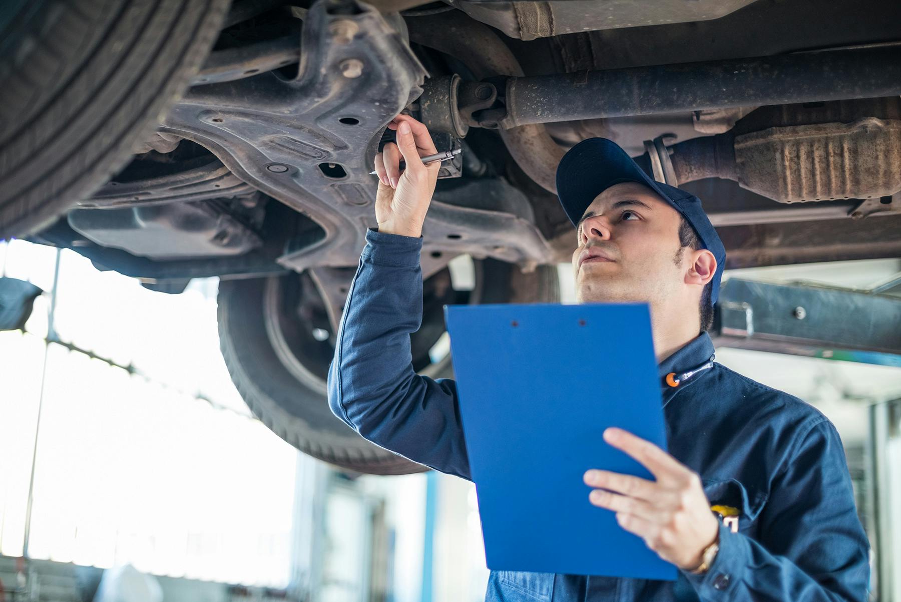 A mechanic inspecting the underbody of a car holding a clipboard