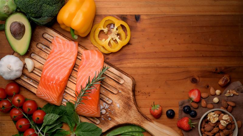 A wooden board with two salmon filets surrounded with vegetables including yellow bell peppers and spinach, which are known immune-supporting foods.