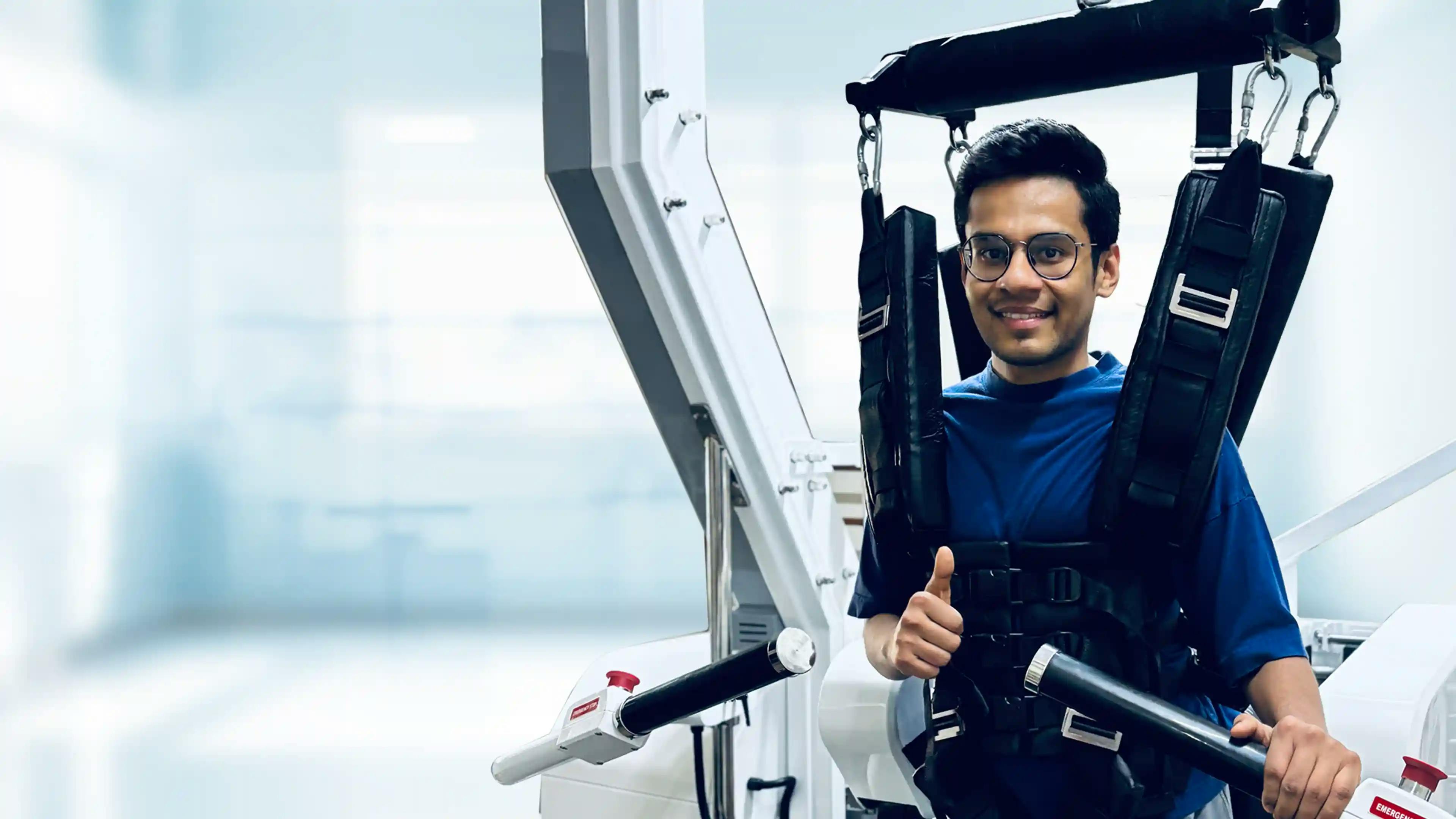 A young man supported in a robotic-assisted gait training device, smiling and giving a thumbs-up during a therapy session.
