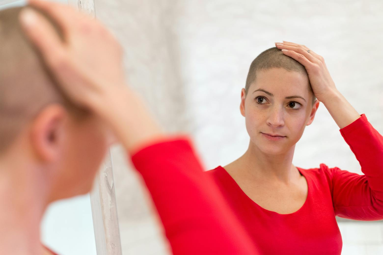 Bald woman looks in the mirror, worried that her cancer chemotherapy treatment will be delayed because of the coronavirus.