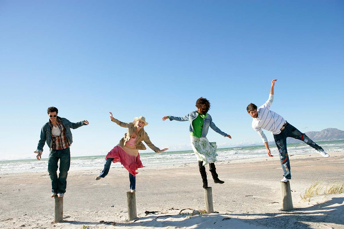 Four diverse people laughing as they struggle to balance on wooden poles at the beach