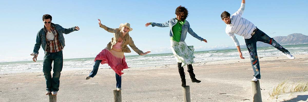 Four diverse people laughing as they struggle to balance on wooden poles at the beach