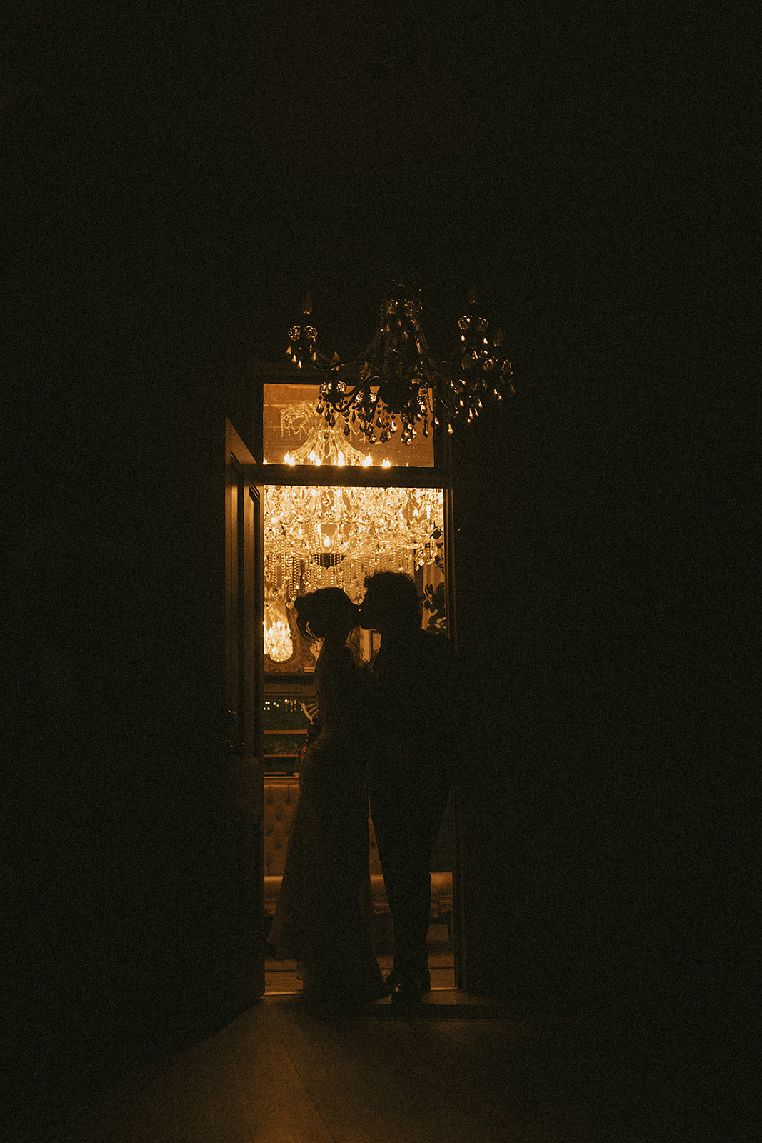 moody chandelier lit photo of a bride and groom sneaking a kiss during their wedding reception