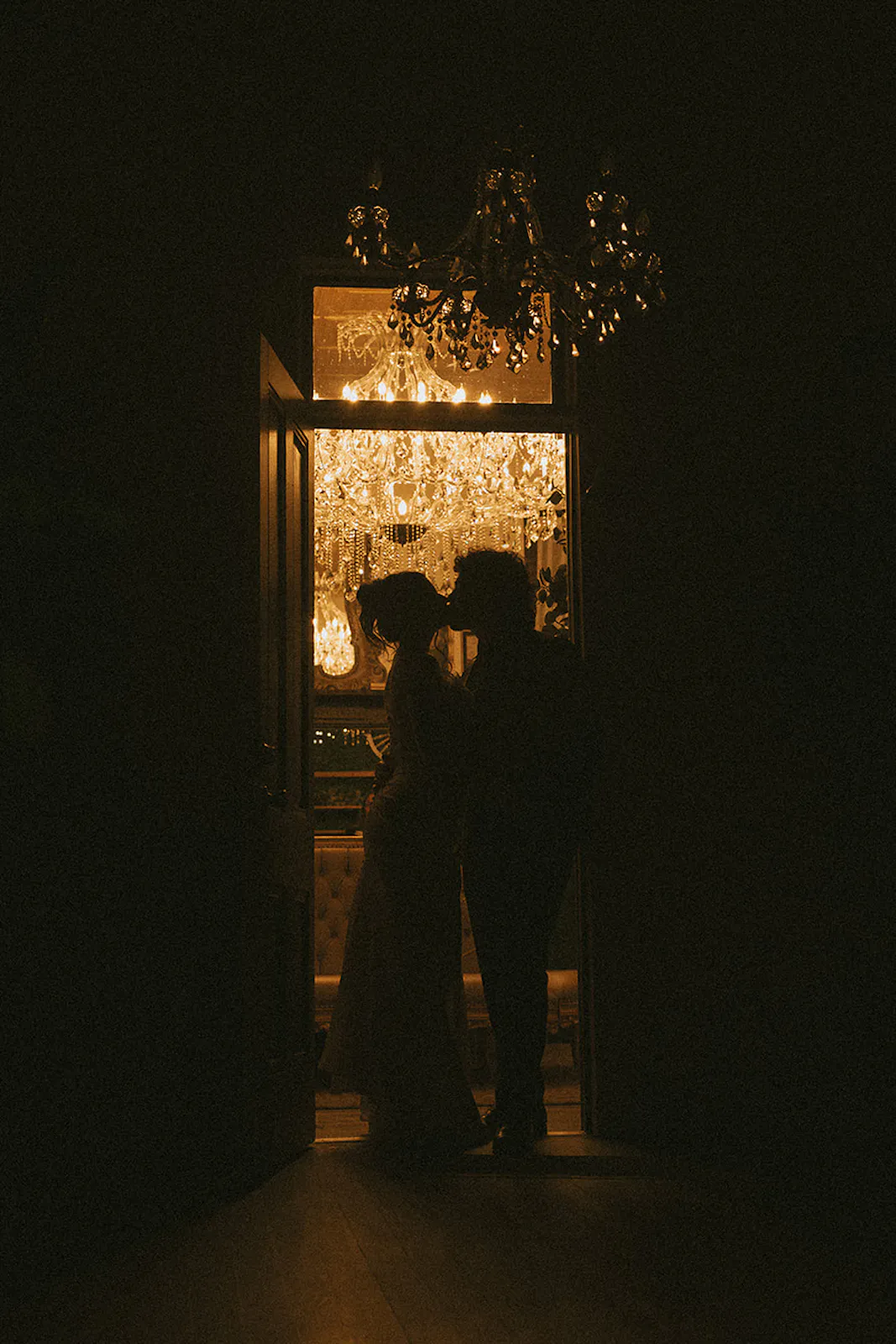 moody chandelier lit photo of a bride and groom sneaking a kiss during their wedding reception