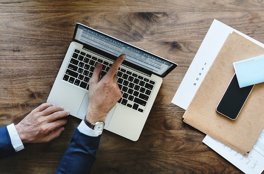 personne montrant l'écran d'un ordinateur portable posé sur une table en bois à côté de documents