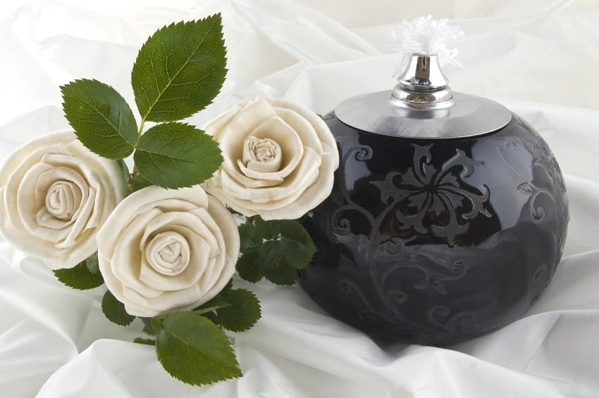 urne funéraire noire posée sur tissu blanc à côté de laquelle sont posées trois roses blanches