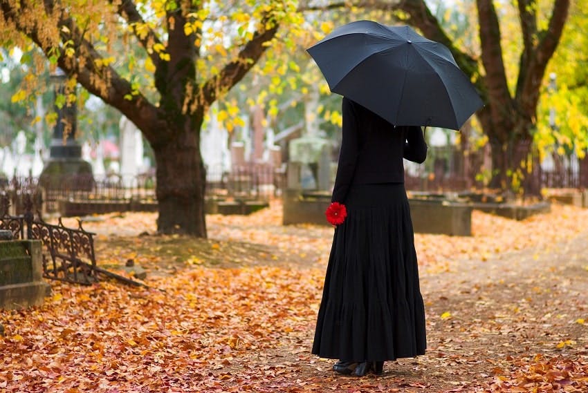 Femme habillée en noir, tenant un parapluie noir ouvert et une fleur rouge dans un cimetière à l'automne
