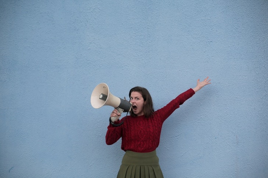 Eine Frau in einem roten Pullover steht mit einem Megaphon vor einer blauen Wand. Die eigene Stimme richtig einzusetzen, ist für Lehrerinnen und Lehrer eine essentielle Fähigkeit. In unserem Beitrag erklären wir dir, wie du deiner Stimme etwas Gutes tun kannst.