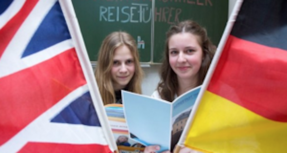 Auf dem Bild sind zwei Mädchen, die zwischen einer britischen und einer deutschen Flagge stehen und einen Reiseführer in der Hand halten. Die Schüler und Schülerinnen des Humanistischen Greifenstein Gymnasiums erstellten einen Reiseführer für die Stadt Thum.