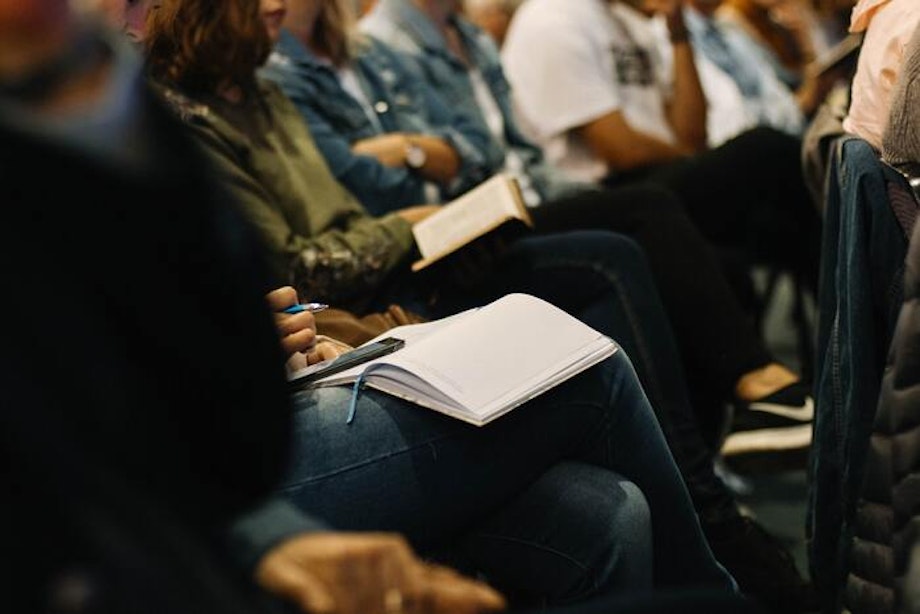 Person sitzt mit aufgeschlagenem Notizbuch in einer Reihe mit anderen Konferenzteilnehmenden