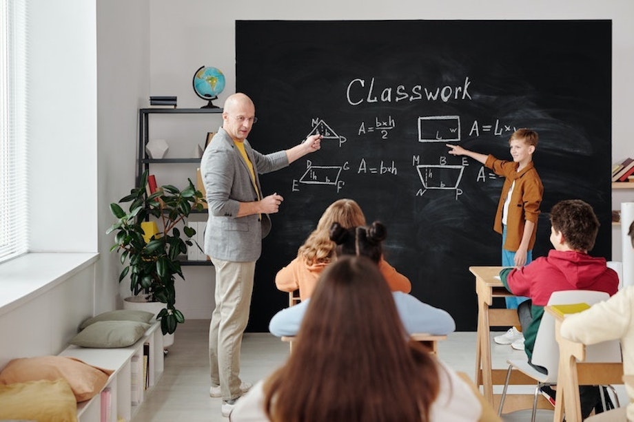 Ein Mathematiklehrer und ein Schüler stehen vor der Tafel und zeigen mit dem Finger auf geometrische Formen, die anderen SchülerInnen hören interessiert zu