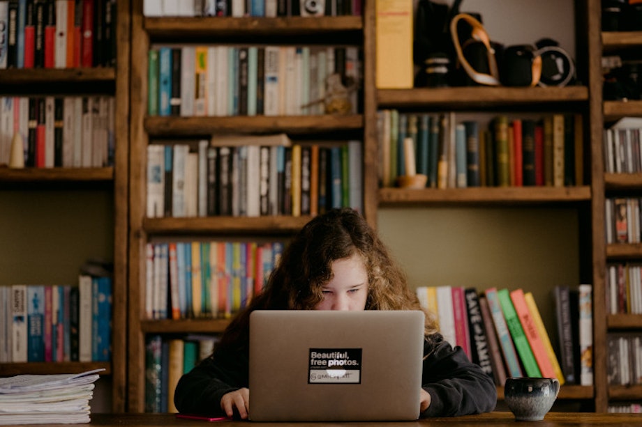 Ein Mädchen sitzt zuhause an einem Laptop vor einem großen Bücherregal. Wer aus einem Haushalt mit hohem Bildungsgrad kommt, hat es beim Homeschooling wesentlich leichter, was soziale Ungleichheit verstärkt.