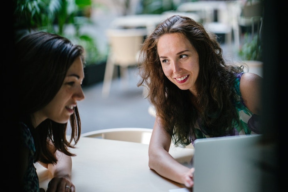 Zwei junge Frauen sitzen an einem Tisch, reden miteinander und schauen auf einen Computerbildschirm.