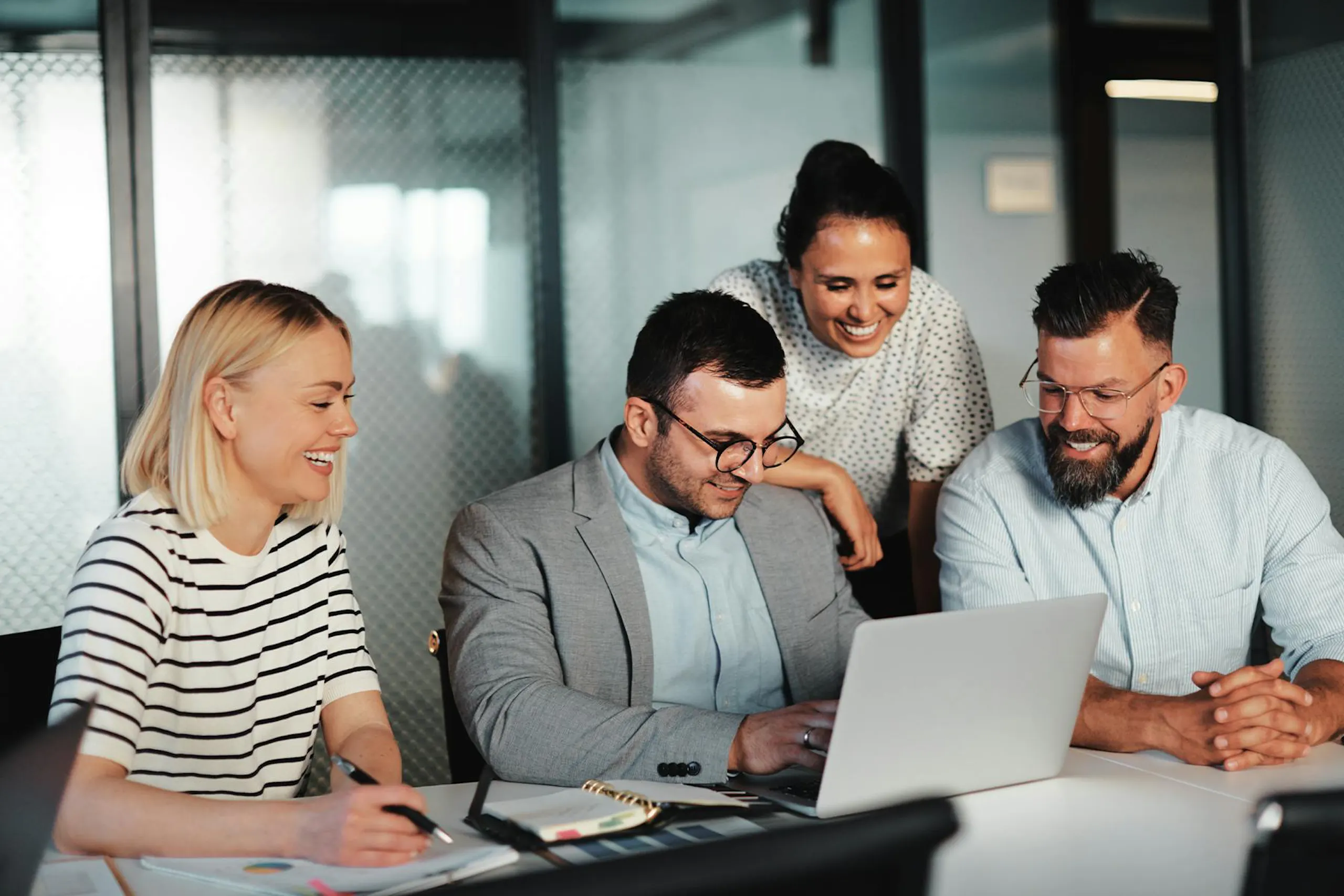 Four people in a meeting room look at a laptop screen while smiling