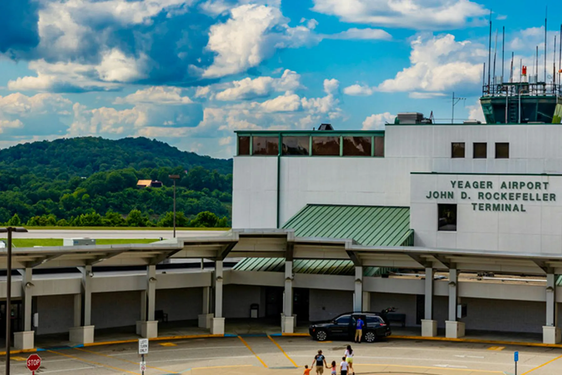 Exterior view of Yeager Airport’s John D. Rockefeller IV Terminal, a white building with green metal roofs and a control tower, set against tree-covered hills and a partly cloudy sky.
