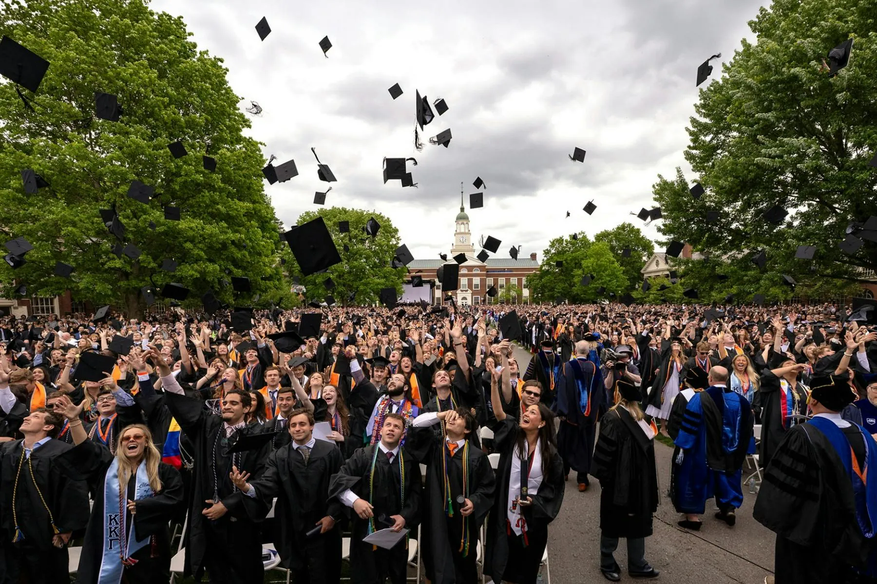 Hundreds of Bucknell University graduates in black caps and gowns celebrate at an outdoor commencement ceremony, tossing their mortarboards into the air as a campus building and large trees frame the scene under a cloudy sky.