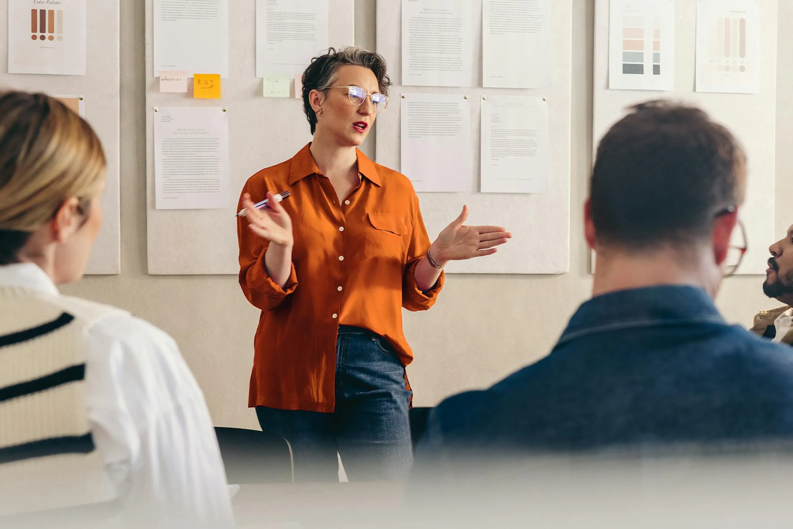 Professional presenting to colleagues during a team meeting, standing in front of a wall displaying reports, design mockups, and notes while discussing ideas and strategy in a collaborative office setting.