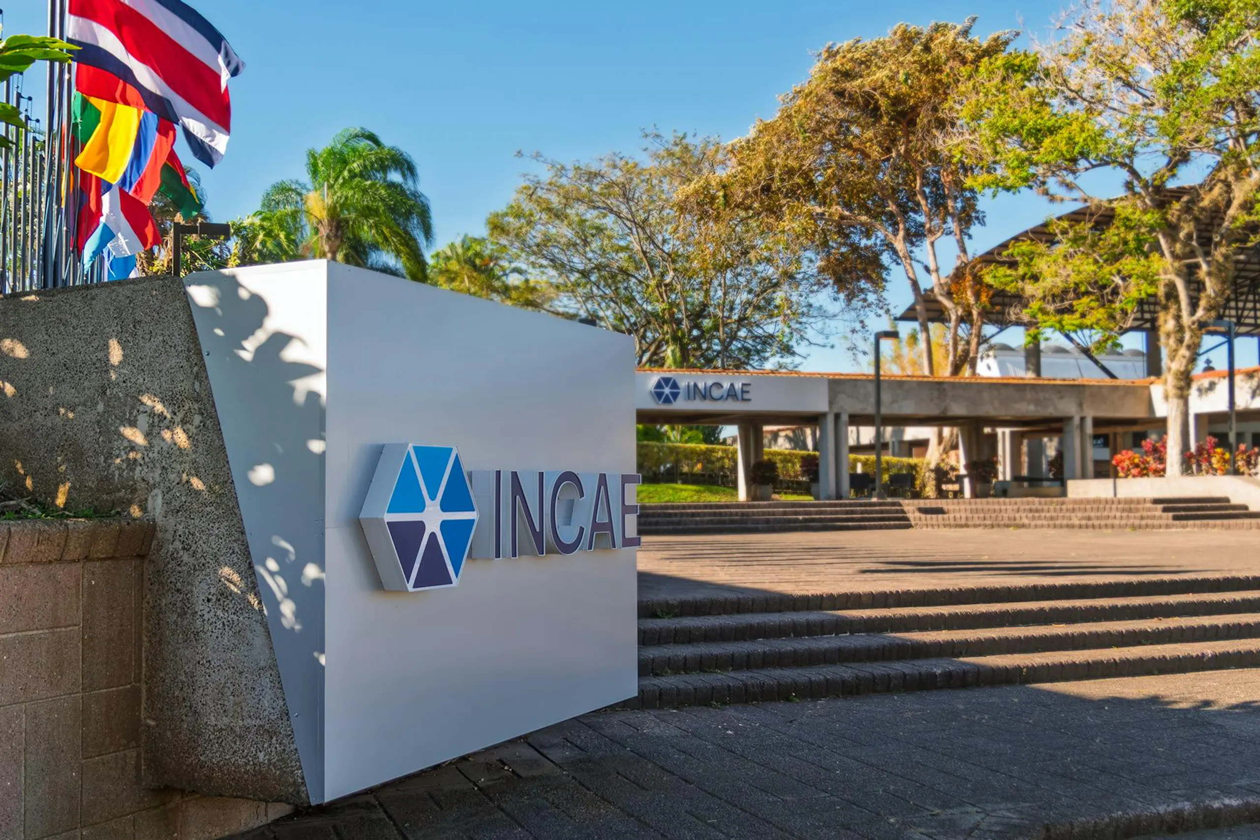 Exterior view of INCAE Business School campus with a large INCAE sign in the foreground, palm trees and flags to the left, and the main building entrance in the background under a clear blue sky.