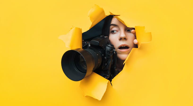 Man sieht eine junge Frau mit einer Kamera mit einem großen Objektiv durch eine gelbe Pappwand luken. Das Bild ist Header für unseren Beitrag zu elektronischen Pressespiegeln.