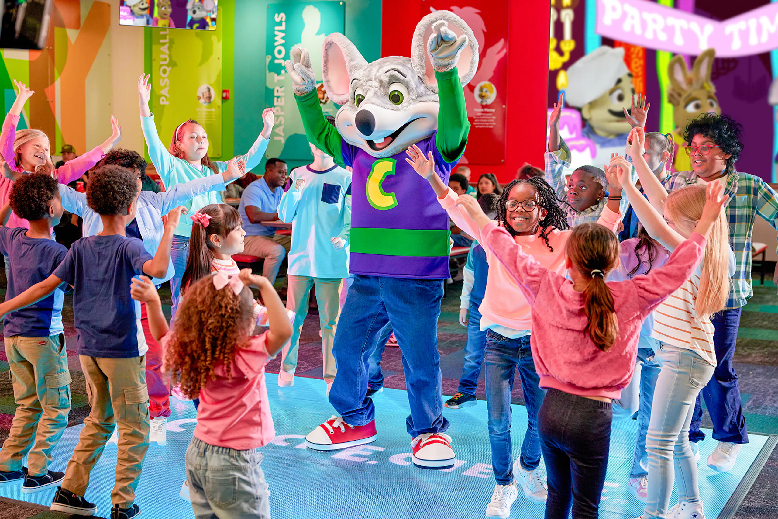 Chuck E. Cheese mascot dancing with a group of children inside a colorful Chuck E. Cheese entertainment center.