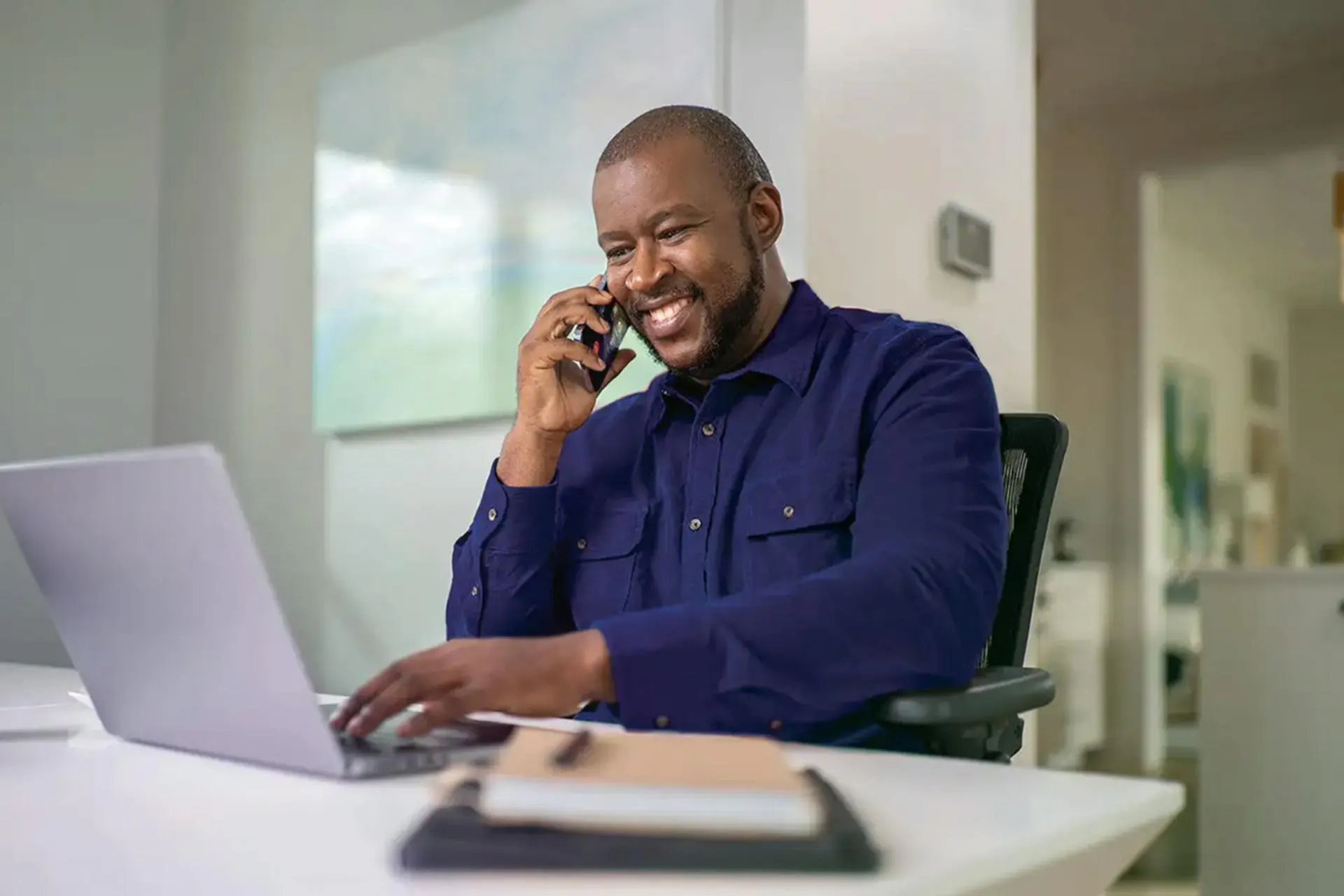 A man sitting in front of a laptop and speaking on a cellphone.