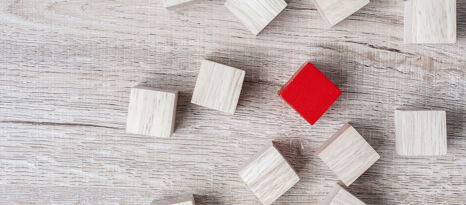 Man sieht einen weiß gestrichenen Holztisch, auf dem viele weiße Holzwürfel zu sehen sind. In der Mitte sticht ein roter Würfel hervor. Dieses Bild ist das Titelbild unseres Beitrags zur Erarbeitung eines USPs / Alleinstellungsmerkmals.