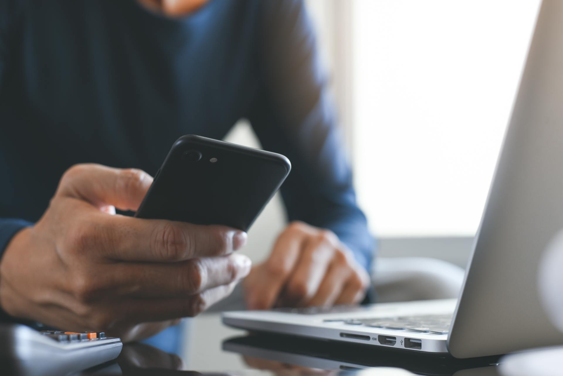 man holding mobile phone with laptop on the desk