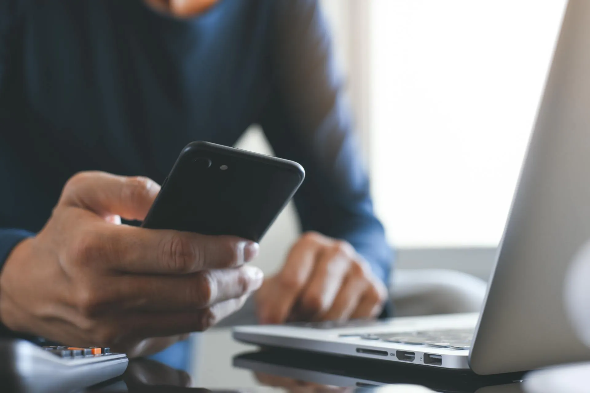 man holding mobile phone with laptop on the desk