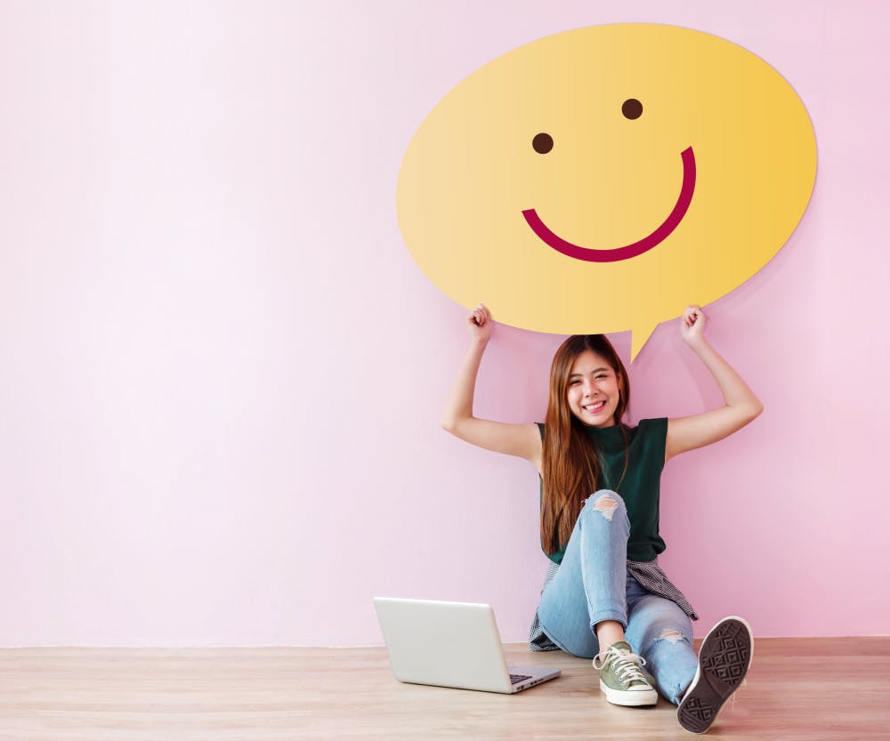 Junge asiatische Frau sitzt mit ihrem Laptop auf dem Boden vor einer pinken Wand und hält einen großen Smiley über ihren Kopf