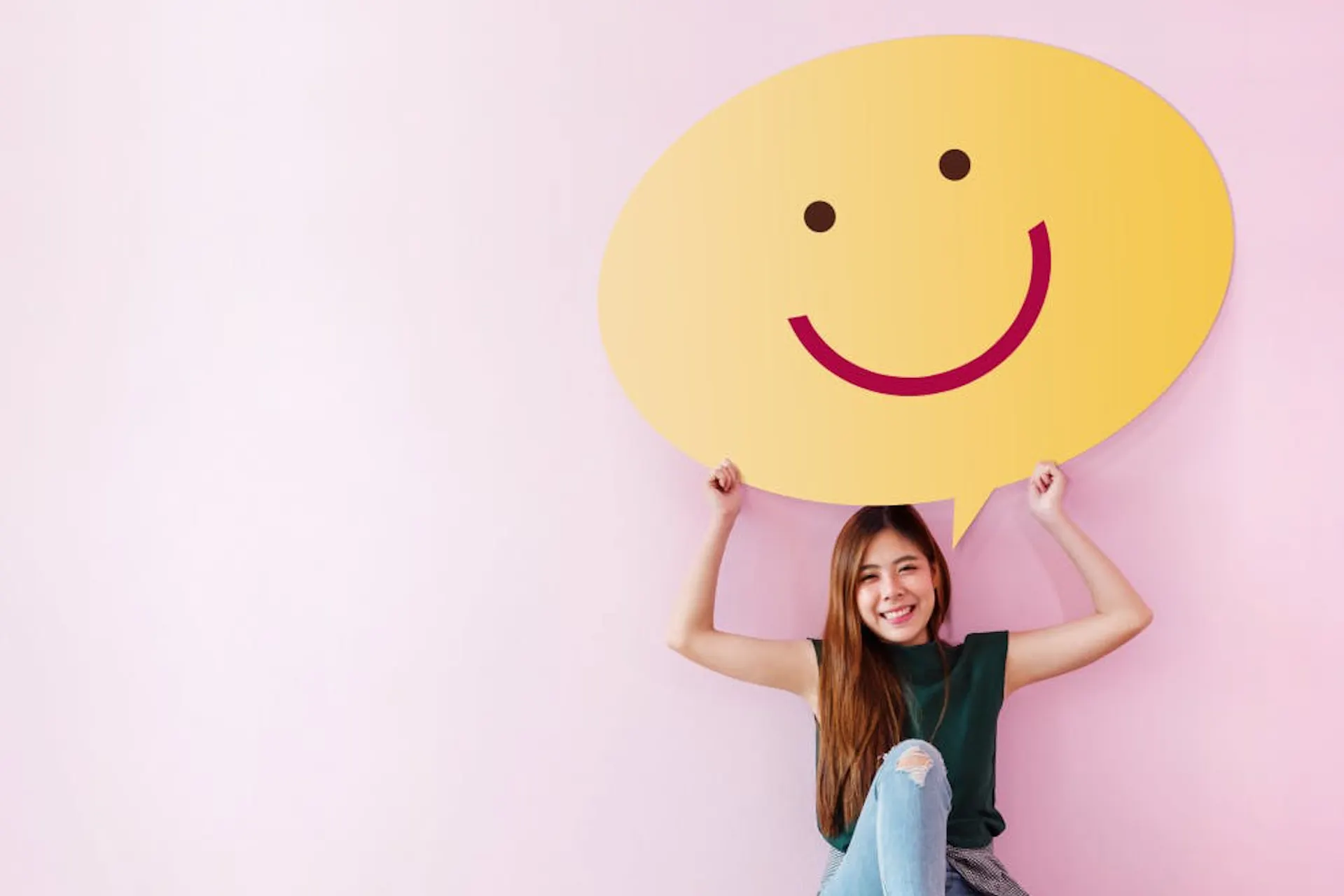Junge asiatische Frau sitzt mit ihrem Laptop auf dem Boden vor einer pinken Wand und hält einen großen Smiley über ihren Kopf
