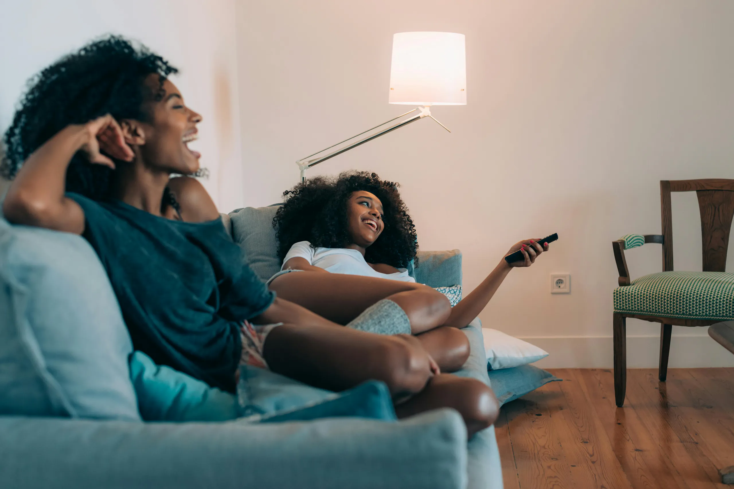 Two women sitting on a softa casually, with one pointing a TV remote control 