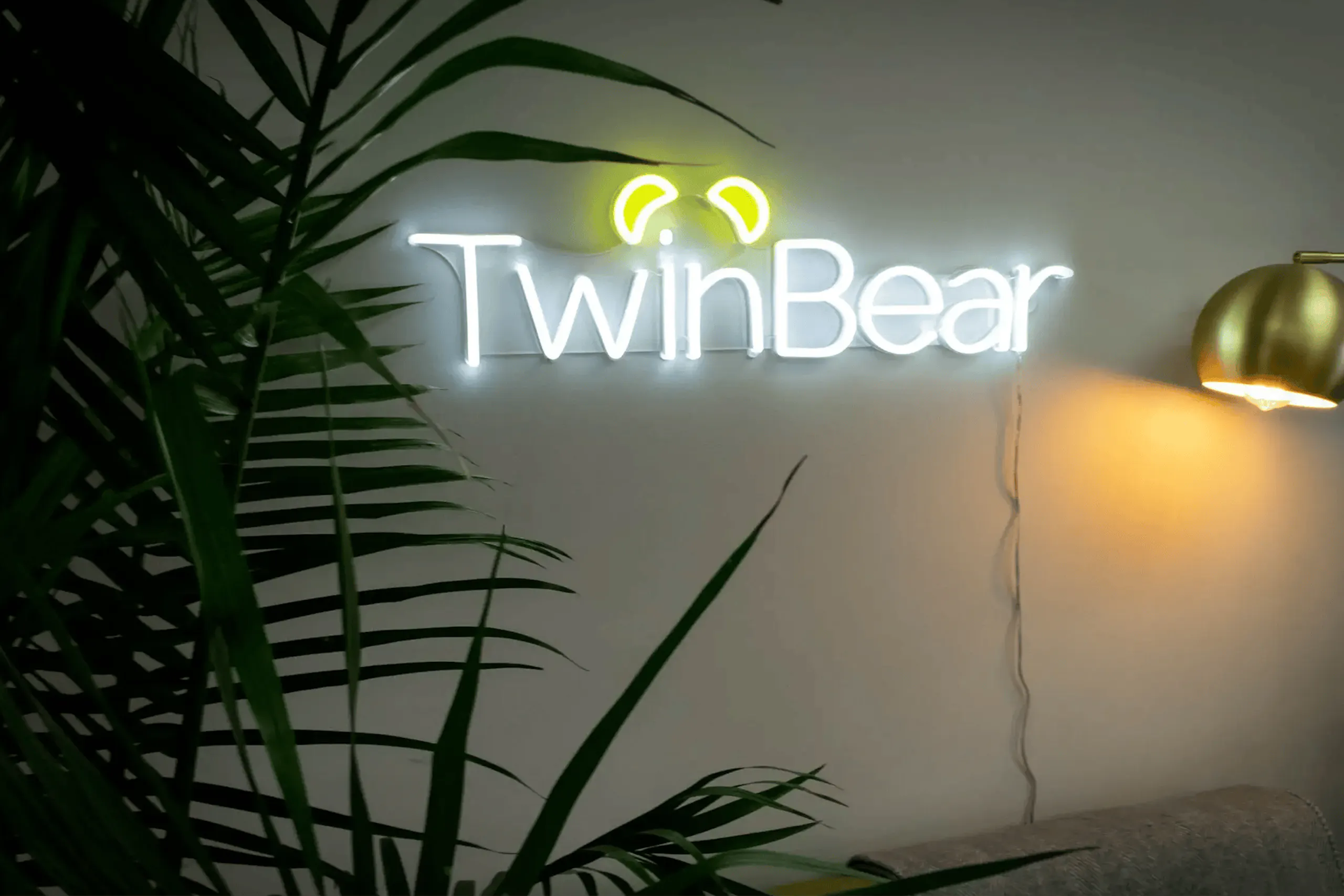 Illuminated TwinBear neon wall sign with stylized bear ears above the logo, mounted on a light-colored wall beside a gold floor lamp and surrounded by green indoor plants.