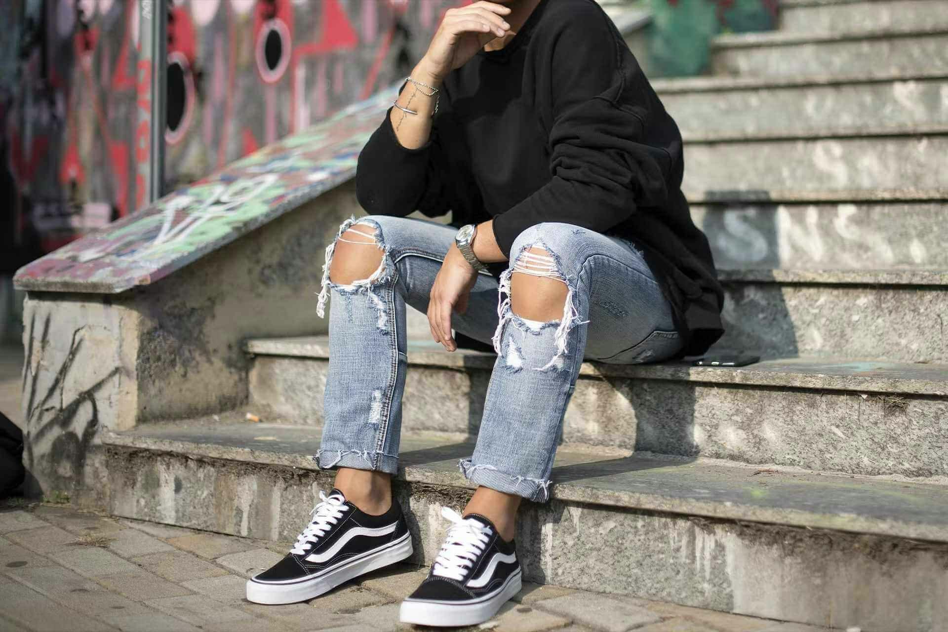 Personne assise sur les marches d'un escalier en extérieur - elle porte un jean bleu troué, un sweat noir et des Vans