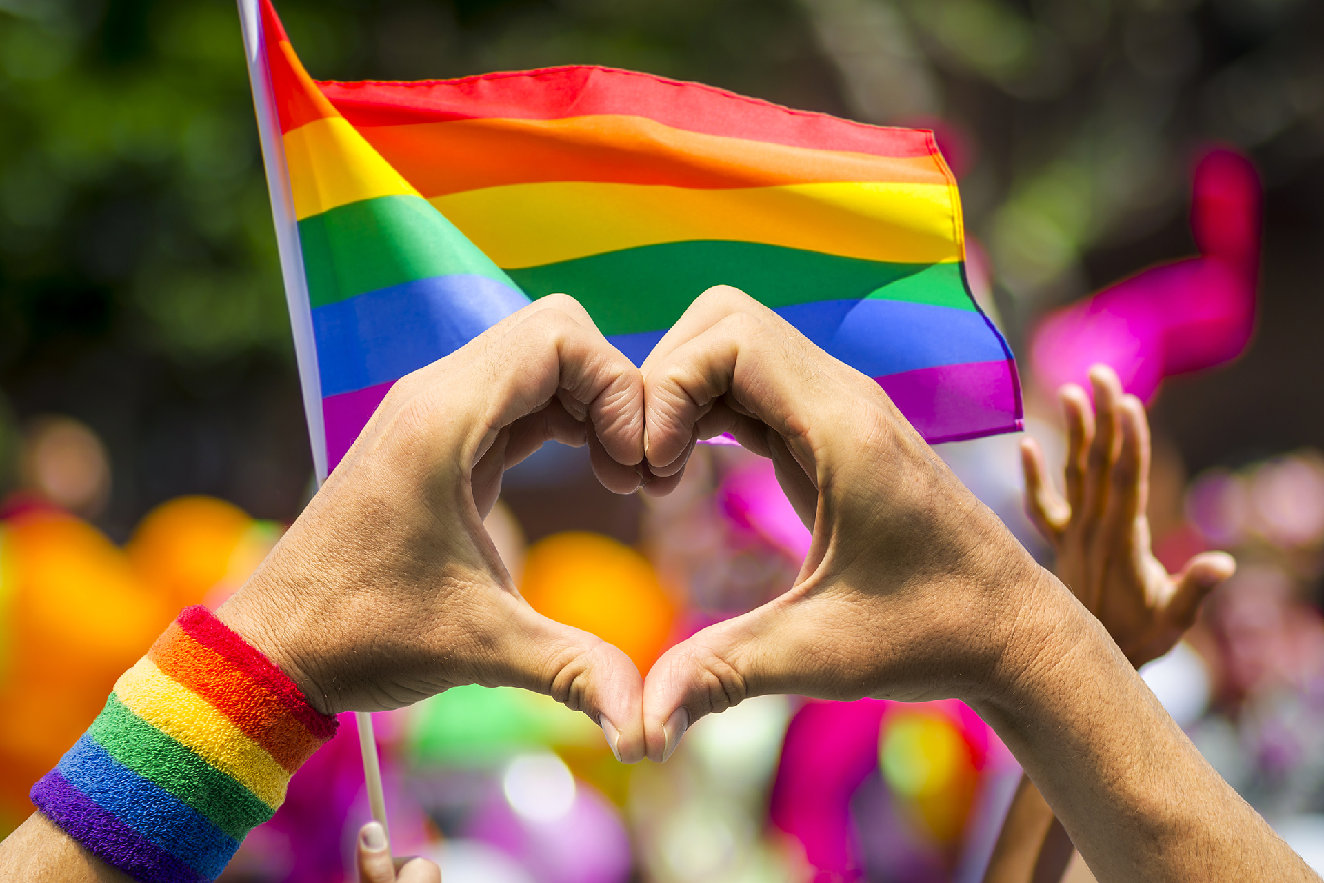 Photo of a rainbow flag during a parade with someone making a heart with his hands in the foreground