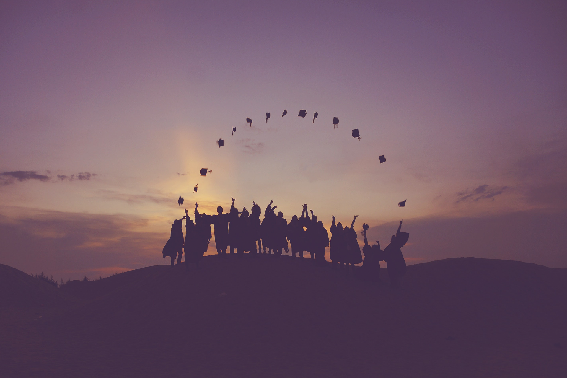 Photo of students throwing their hats into the air while standing on a hill in the sunset.